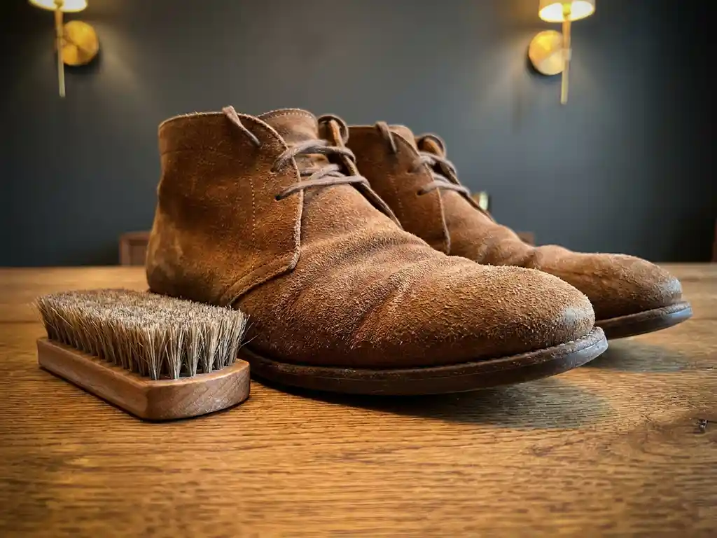 Versleten suède schoenen in cognac en taupe op eiken houten ondergrond, met een natuurlijke borstel voor suède onderhoud.